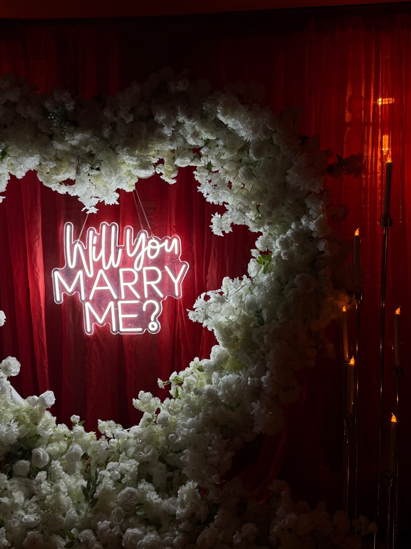 White floral heart with a neon proposal sign against red drapes