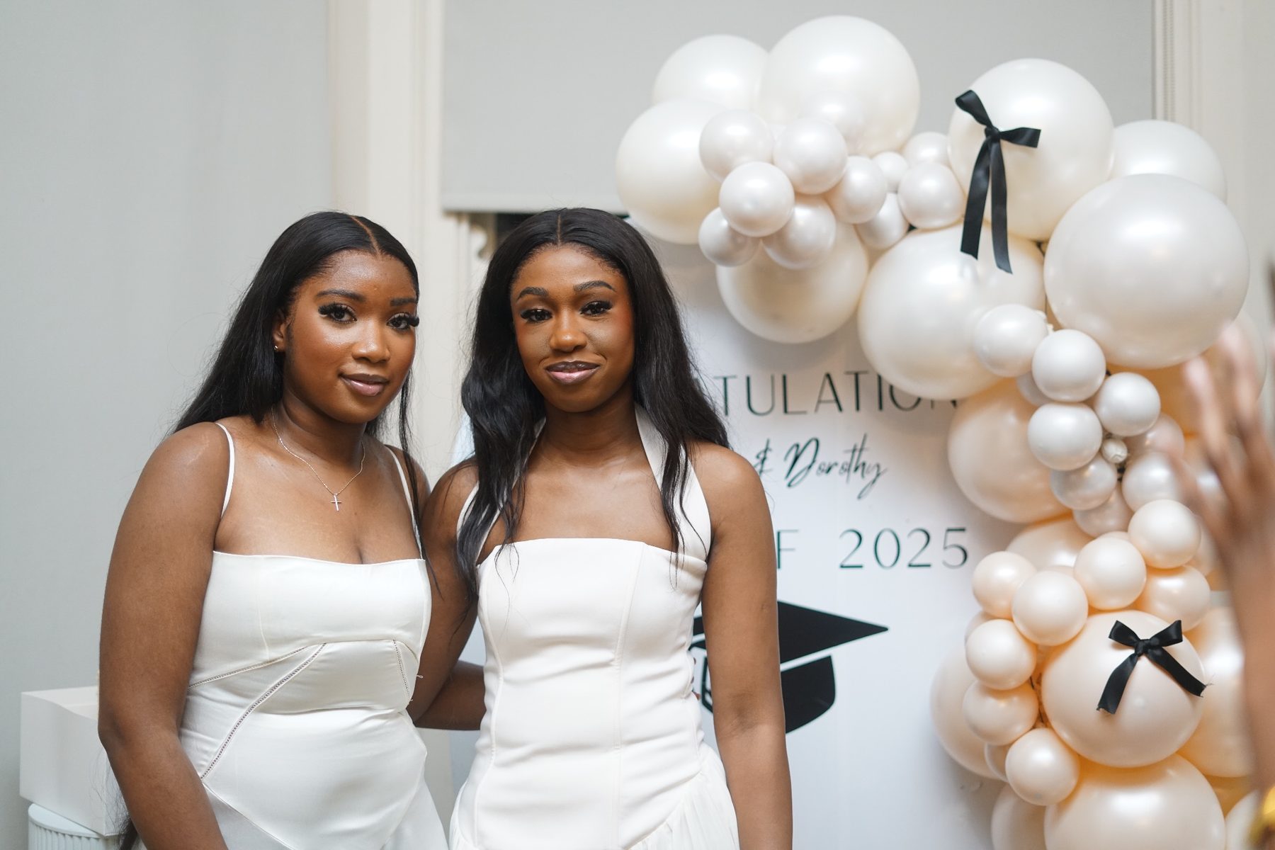 Two graduates in white in front of a pearl balloon backdrop