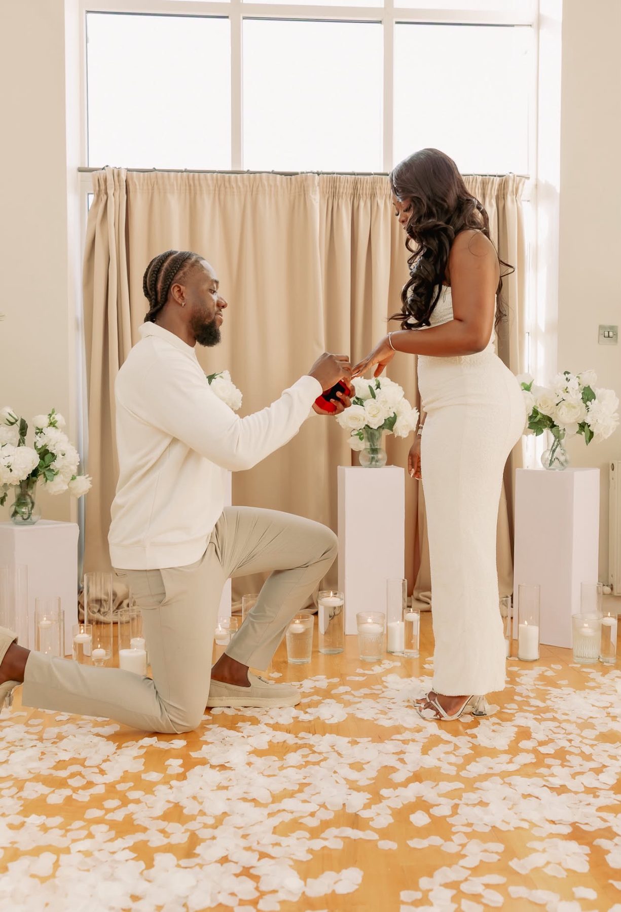 A man kneels to propose in a room styled with white roses and candles